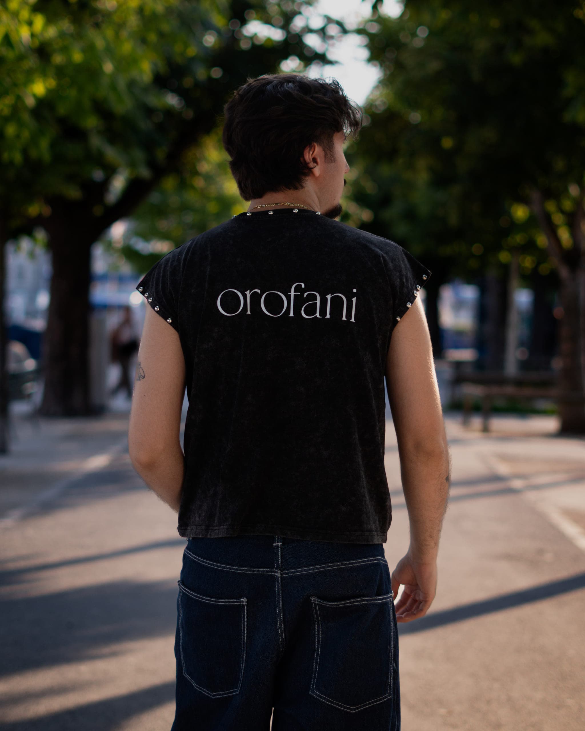 Man seen from behind in a sleeveless black shirt with lettering on the back, on a tree-lined promenade