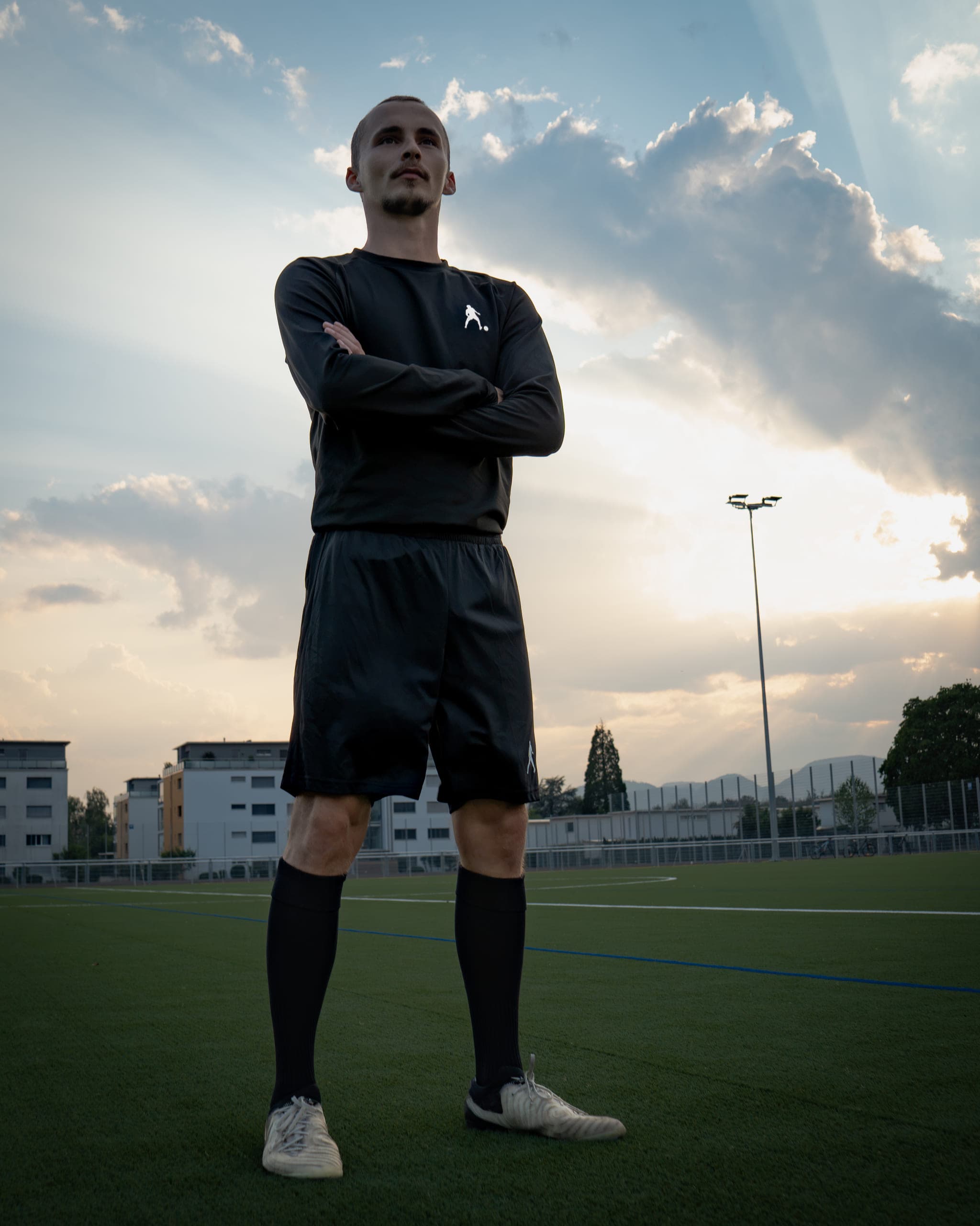 Man in black sportswear standing with crossed arms on a football pitch under a dramatic evening sky