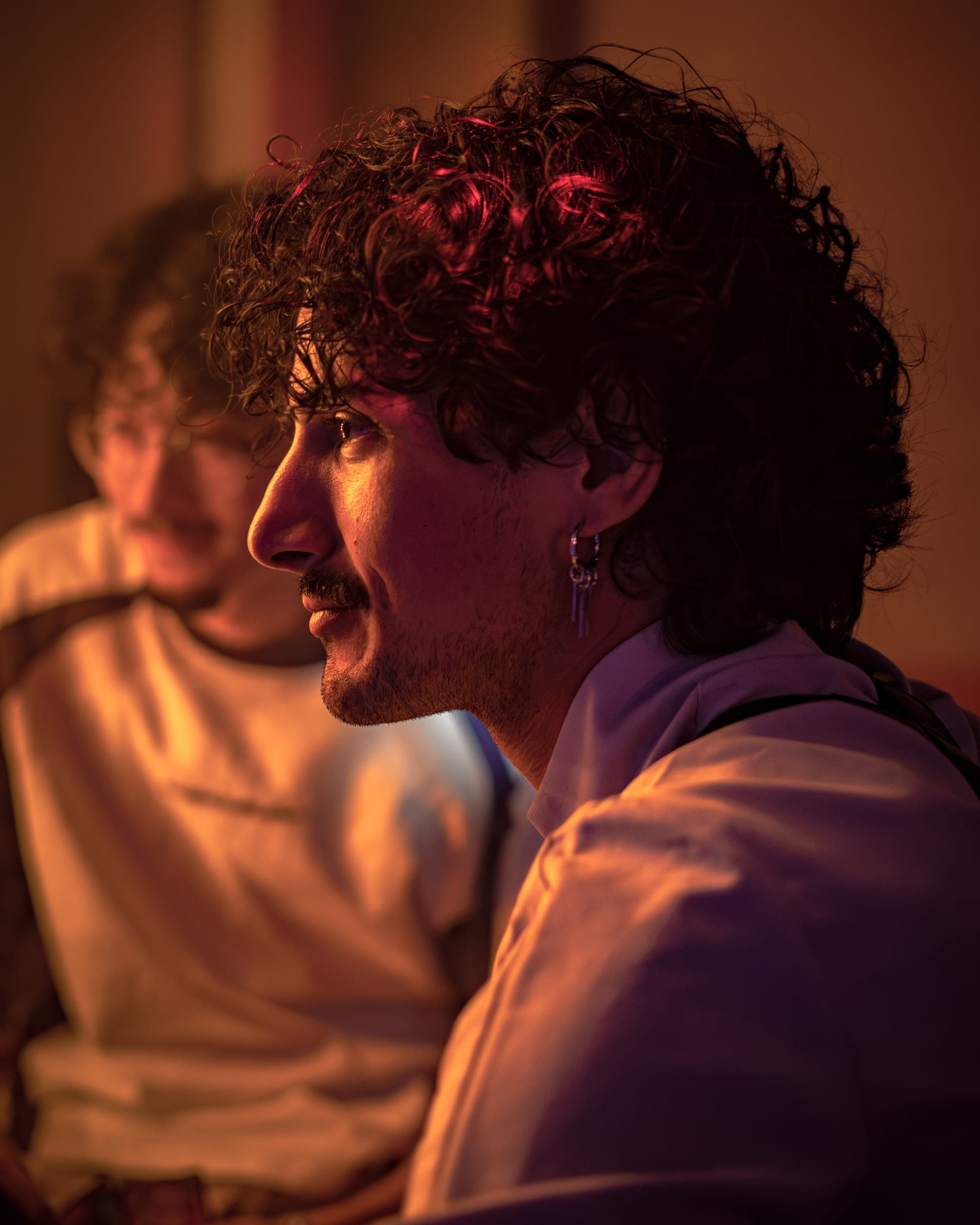 Profile of a man with curly hair and an earring in warm red and orange light, a friend blurred beside him