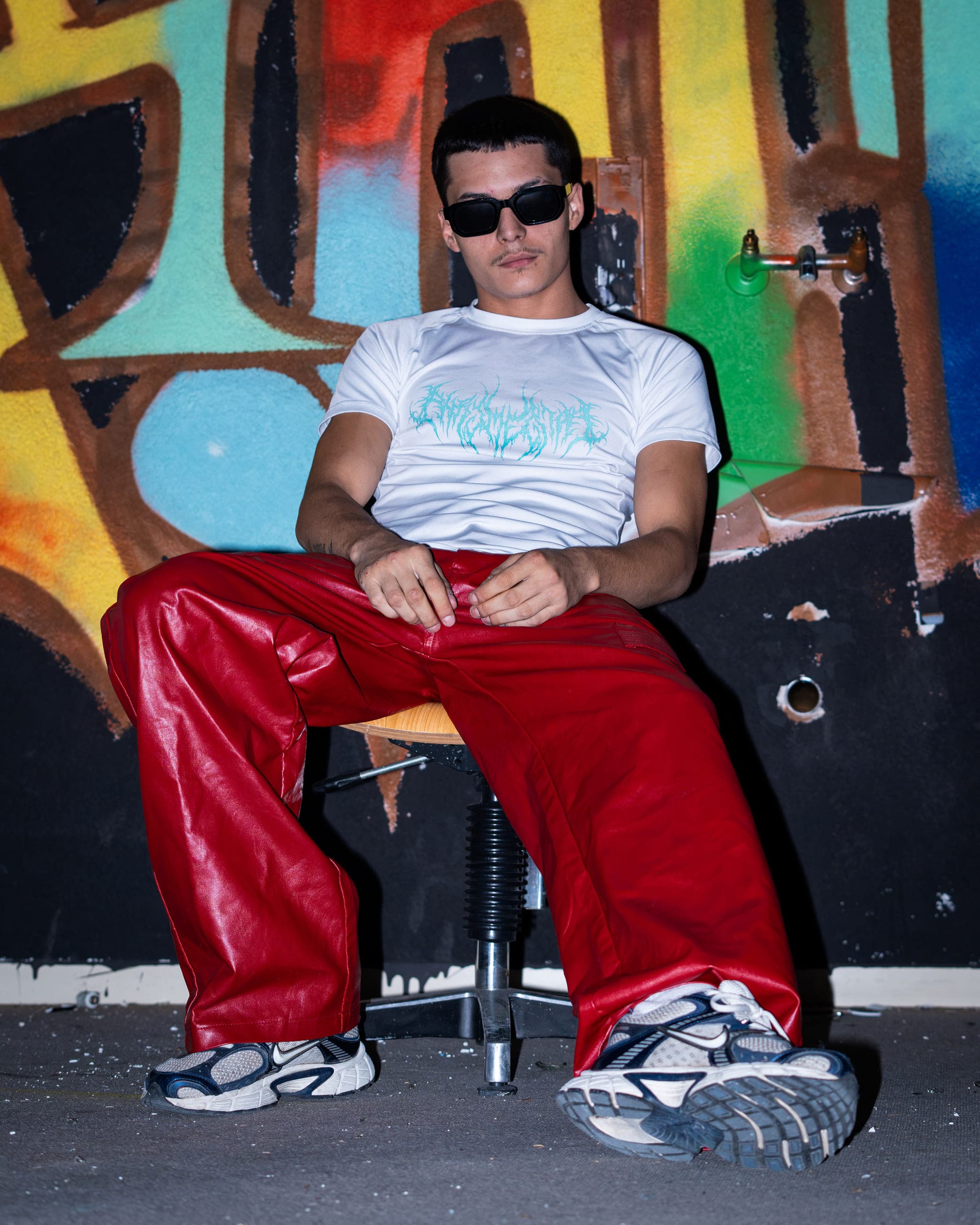 Man in sunglasses, white T-shirt and glossy red trousers sitting on a stool in front of a colourful graffiti wall
